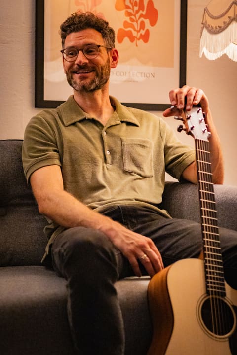 Jochen sitzt lachend mit Gitarre auf dem Sofa.